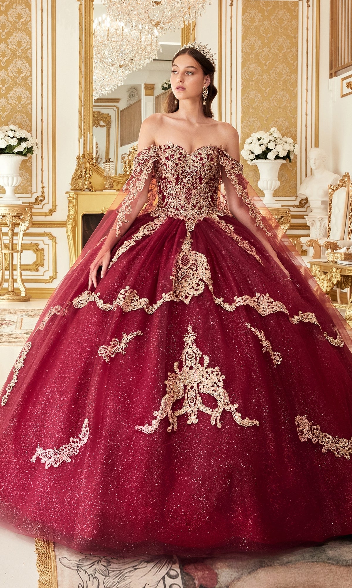 A woman wears the Ladivine 15711 Quinceañera Dress with gold embroidery—an off-shoulder burgundy ball gown adorned with glitter tulle—in an ornate, gold-accented room featuring chandeliers, mirrors, and white floral arrangements.